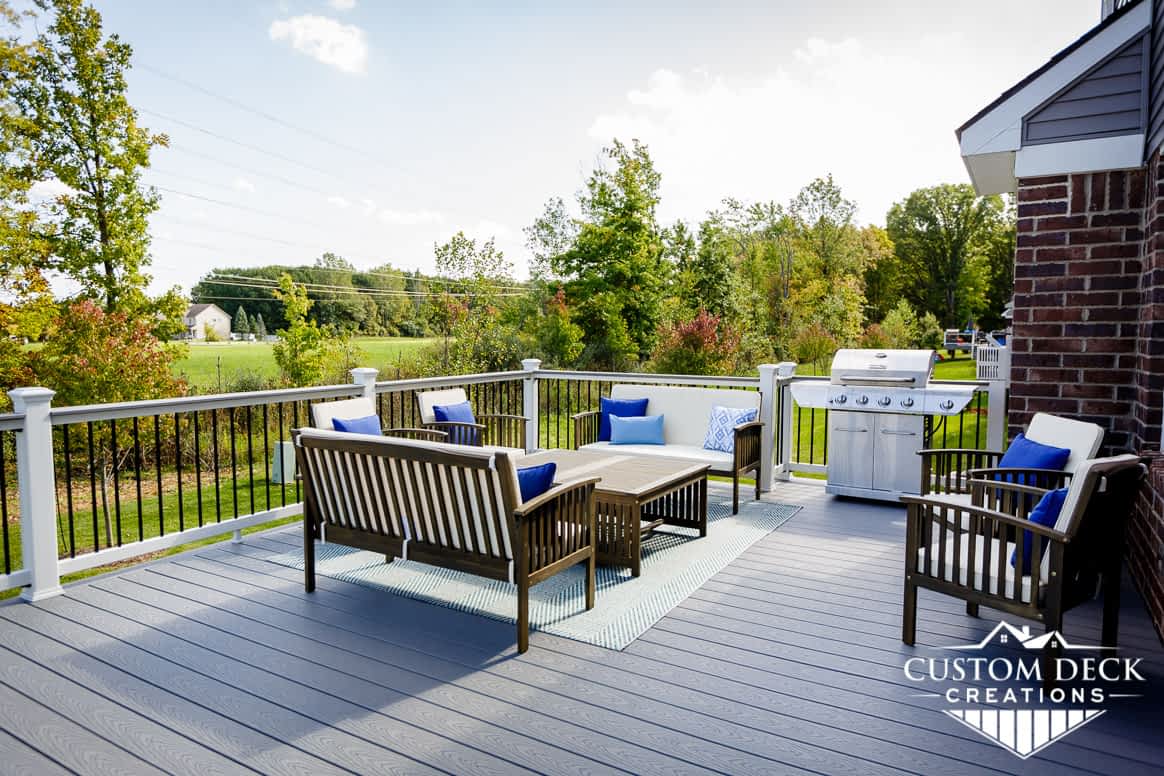 Grey deck with brown patio furniture, and a grill, in a backyard behind a home