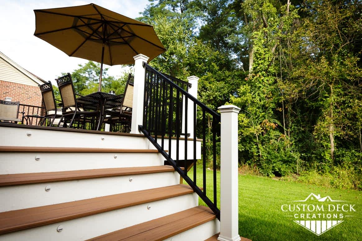 Detailed view of composite stairs of a backyard deck showing white and black railing, stair lights, and shade umbrella on a sunny day