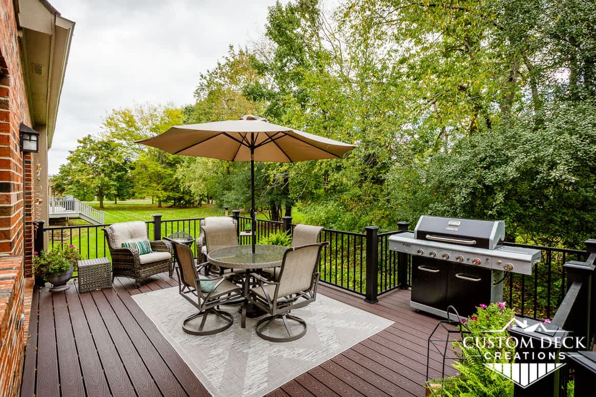 Brown backyard view on top of a deck showing a grill, patio furniture, shade umbrella, and lounge chairs