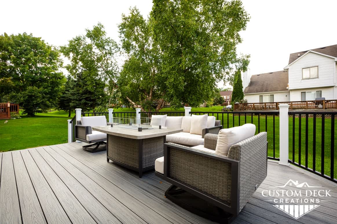 View of a home's backyard from on top of a grey composite deck with black and white railing, shown with gas fire pit and seating area