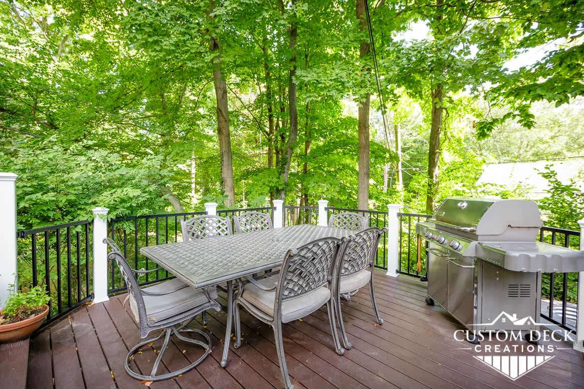 Patio table, chairs, and a grill sitting on a brown backyard deck surrounded by trees
